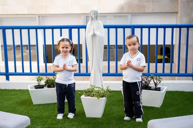 Dos estudiantes en uniforme escolar realizan ejercicio de meditación frente a estatua religiosa en zona verde del colegio
