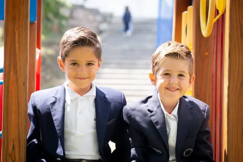 Dos estudiantes con uniforme escolar posando juntos en la entrada del colegio, sonriendo hacia la cámara